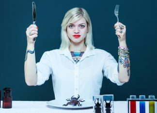 Sind das die 7 Foodtrends von morgen? Eine junge, tättowierte Frau, die Messer und Gabel in die Luft hält. Vor ihr Reagenzgläser und Käfer auf dem Teller