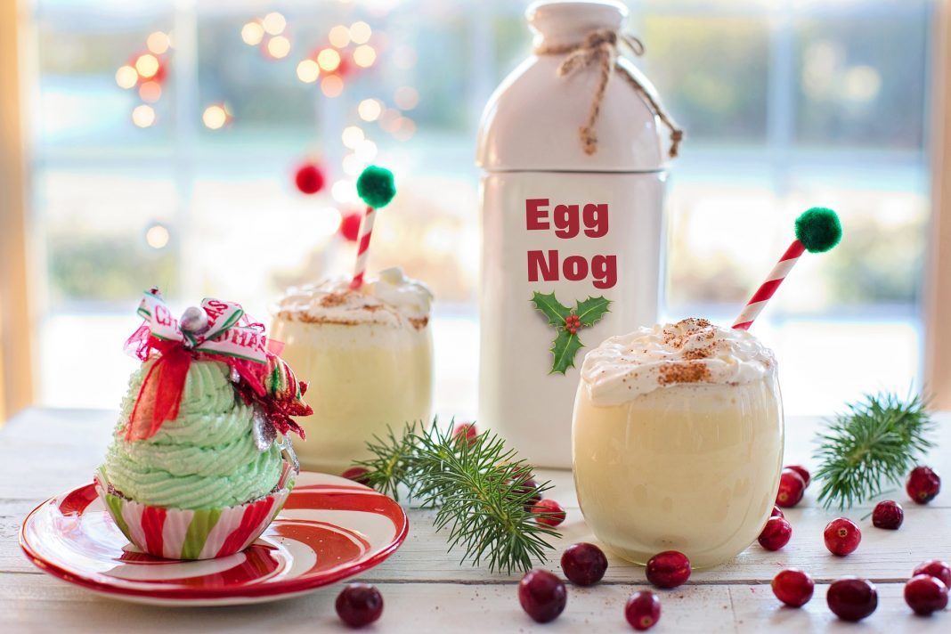 Eine weie Flasche mit der aufschrift "Egg Nog", zwei mit eierlikör gefüllte Gläser und ein grüner Cupcake auf einem weißen Tisch mit roten Beeren und grünen Tannenzweigen.