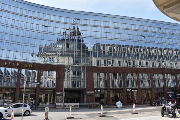 Eine sechsstöckige Glasfassade, in dem sich ein Gebäude mit Turm spiegelt.