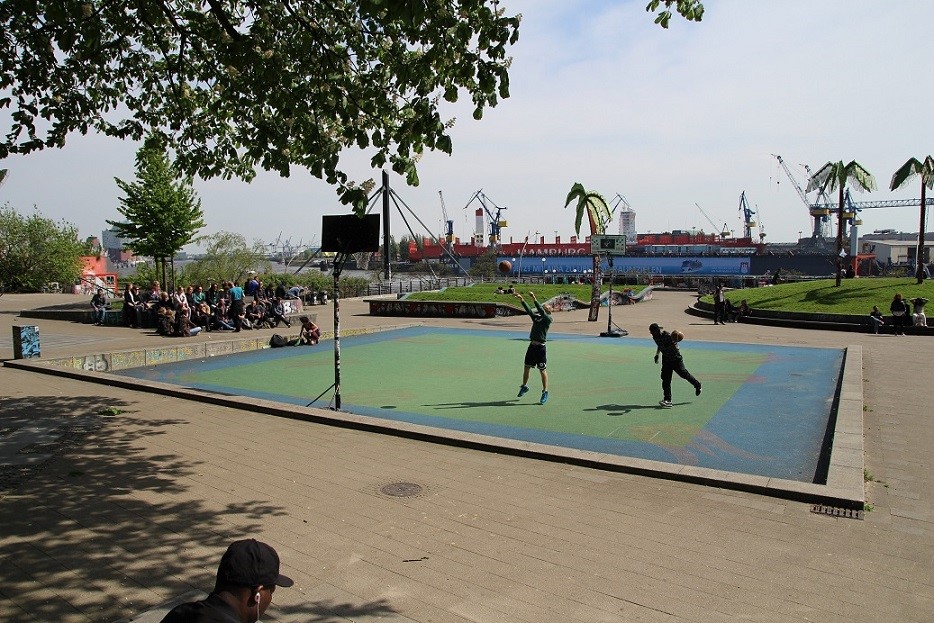 Bild des Park Fiction. Die Sonne scheint, auf dem Basketballplatz wird gespielt, während eine Gruppe von Menschen vom Rand aus zuschaut.