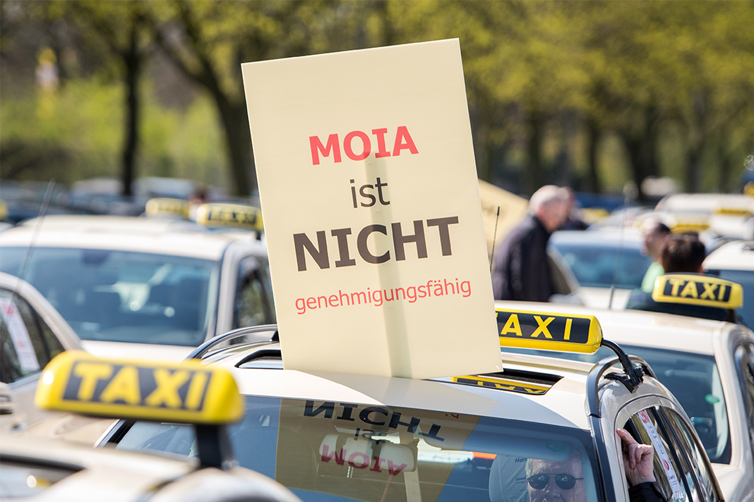 Taxis stehen aufgereiht. Ein Taxifahrer hält aus seinem Auto ein Schild mit der Aufschrift "Moia ist nicht genehmigungsfähig".
