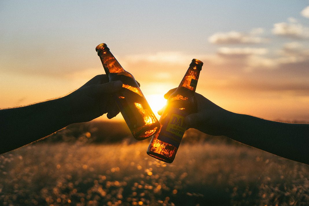 Zwei Hände mit Bierflaschen stoßen vor einem Sonnenuntergang an.