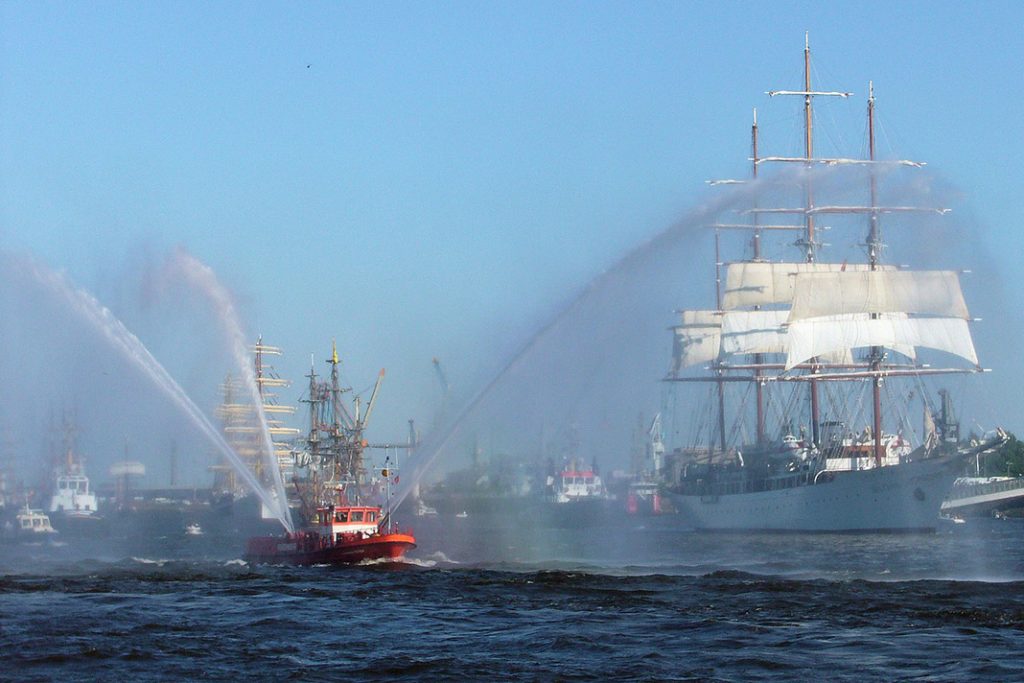 Ein Feuerwehrboot, Ein Viermaster und weitere Schiffe im Hintergrund bei der Auslaufparade