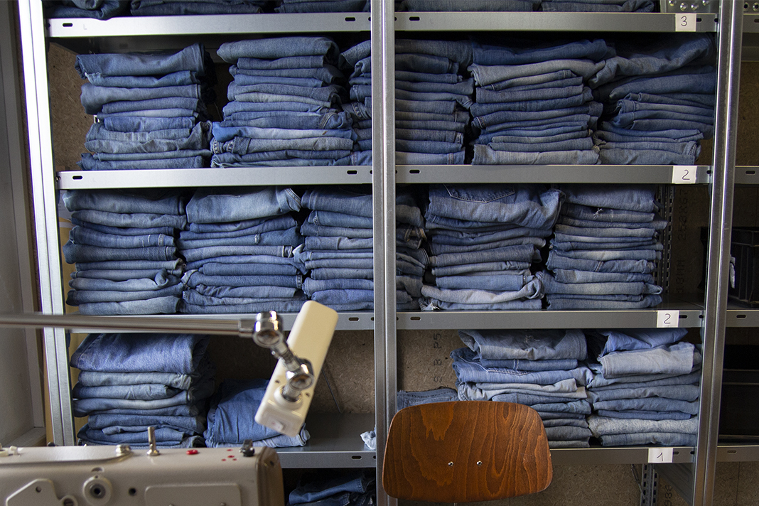Gestapelte blaue Jeans im Regal, davor eine Nähmaschine