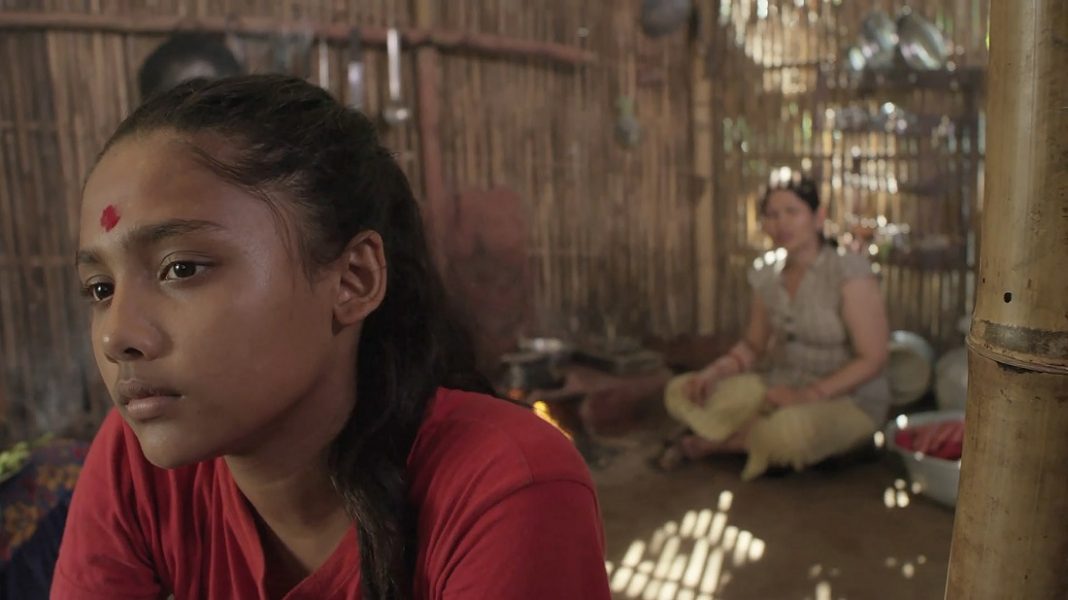 Ein hinduistisches Mädchen sitzt in einer Bambushütte, im Hintergrund unscharf die Mutter