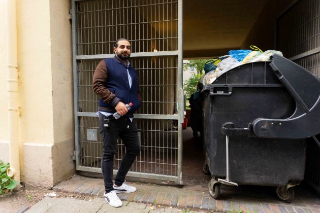 Ali Hakim, der Regisseur des Films "Bonnie &Bonnie", steht vor einer Toreinfahrt, in der Müllcontainer stehen, aus denen die Müllsäcke oben herausgucken.