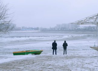 Zwischen Moral und Erfolg Zwei Männer sitzen am Strand in einem gestrandeten Boot und reden miteinander, während es schneit.