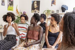 Sechs Frauen mit Afrohaaren, die an einem Haarworkshop von Abina teilnehmen. Sie sitzen in zwei Reihen. Eine Teilnehmerin spricht, die Köpfe der anderen sind ihr zugewandt.