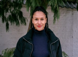 Black Power Germany Black Power Germany Aktivistin Charlotte Nzimiro steht vor einer weißen Mauer in Planten un Blomen. Im Hintergrund ist ein Nadelbaum zu sehen.