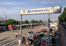 Schoko Jonny, Jessy und Herr Peters: Drei Fischmarkt-Originale feiern Wiedereröffnung Eingang zum Fischmarkt Hamburg an einem sonnigen Tag.