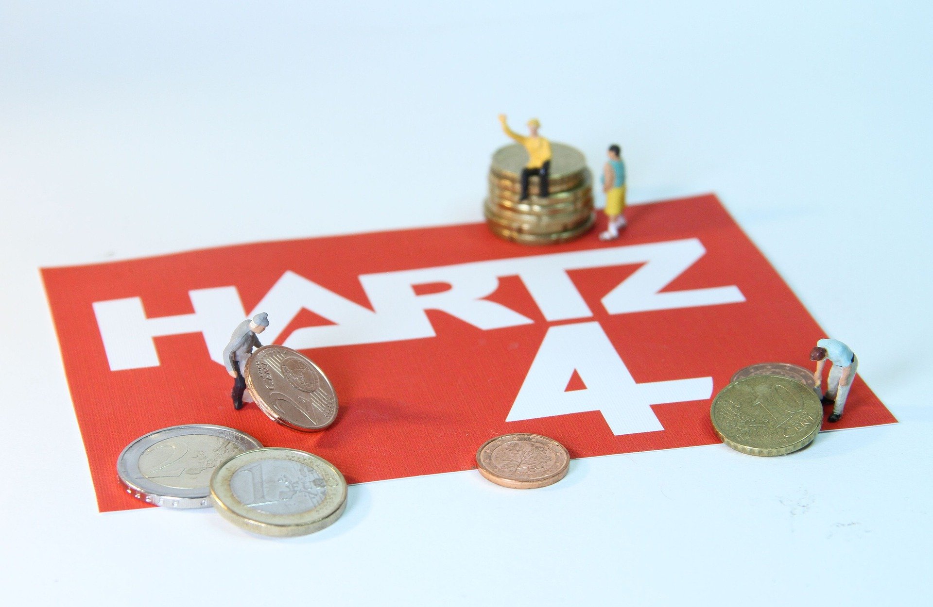 Miniaturfiguren spielen mit Euromünzen. Zwei Figuren heben eine Centmünze hoch, eine sitzt auf einem Stapel Münzen. Auf dem roten Boden unter den Münzen steht „Hartz 4“. Das Bild steht symbolisch für Menschen, die Sozialhilfe beziehen.