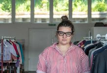 Одяг для нормальності Mila Yazvinska, Projektmanagerin im Help Store, trägt eine schwarze Brille. Sie hat ihre braunen Haare zu einem Dutt gebunden. Sie trägt eine rot weiß gestreifte Bluse und steht zwischen Kleiderstangen.