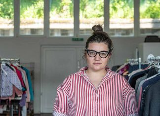 Ein Kleidungsstück Normalität Mila Yazvinska, Projektmanagerin im Help Store, trägt eine schwarze Brille. Sie hat ihre braunen Haare zu einem Dutt gebunden. Sie trägt eine rot weiß gestreifte Bluse und steht zwischen Kleiderstangen.