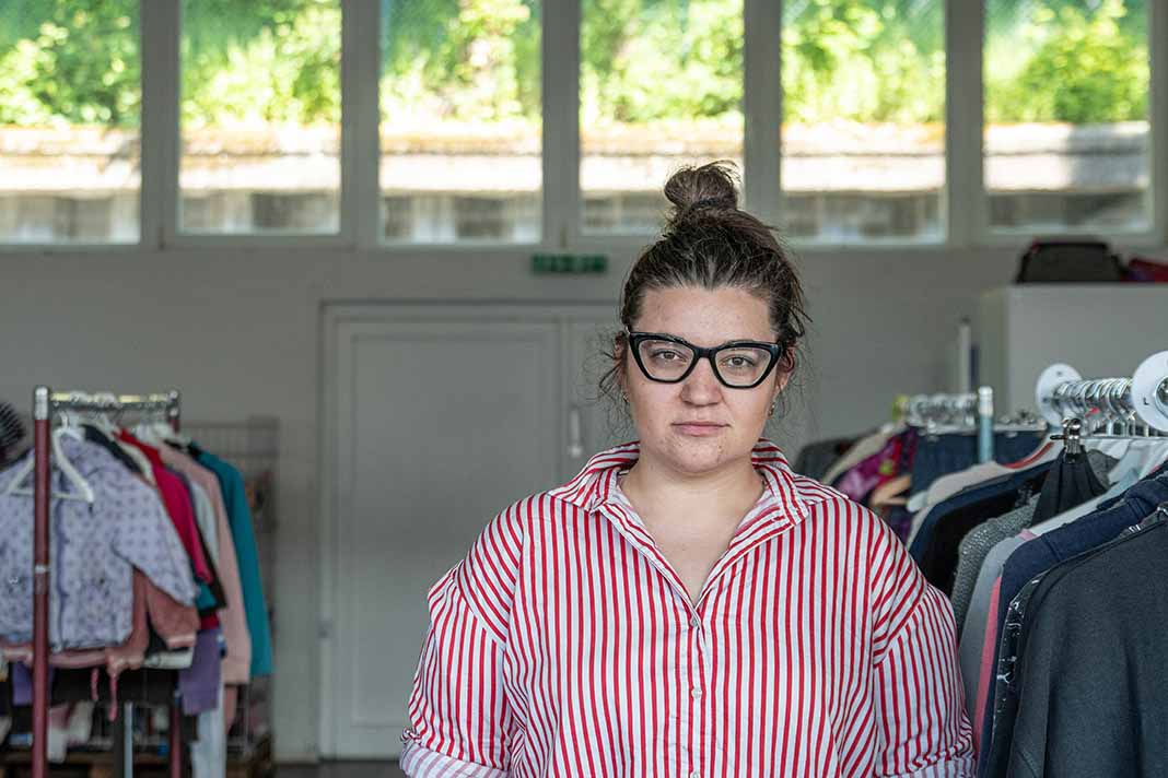Mila Yazvinska, Projektmanagerin im Help Store, trägt eine schwarze Brille. Sie hat ihre braunen Haare zu einem Dutt gebunden. Sie trägt eine rot weiß gestreifte Bluse und steht zwischen Kleiderstangen.