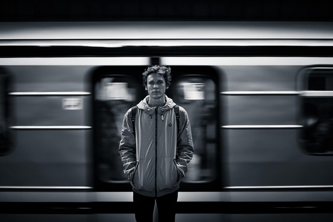 Ein junger Mann steht vor einem Zug. Er dreht ihm den Rücken zu und schaut direkt in dei Kamera, während der Zug vorbeizieht