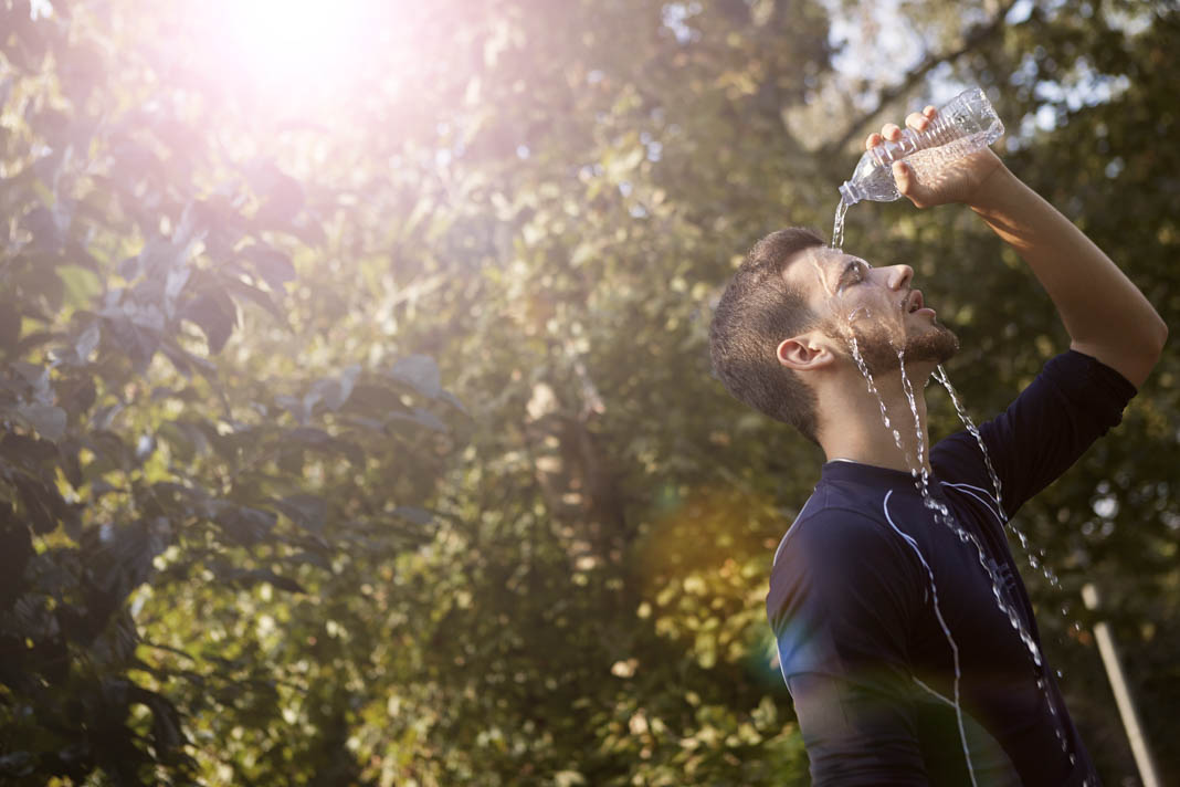 Ein junger Mann gießt sich Wasser aus einer durchsichtigen Flasche auf den Kopf. Es scheint die Sonne.