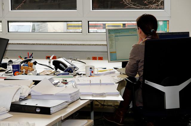 Arbeitslos Hamburg: Eine Frau mit dunklen Haaren sitzt an einem Schreibtisch und arbeitet am Computer. Auf dem Schreibtisch herrscht Chaos. Es liegen Papiere, Ordner und andere Arbeitsmaterialien herum.