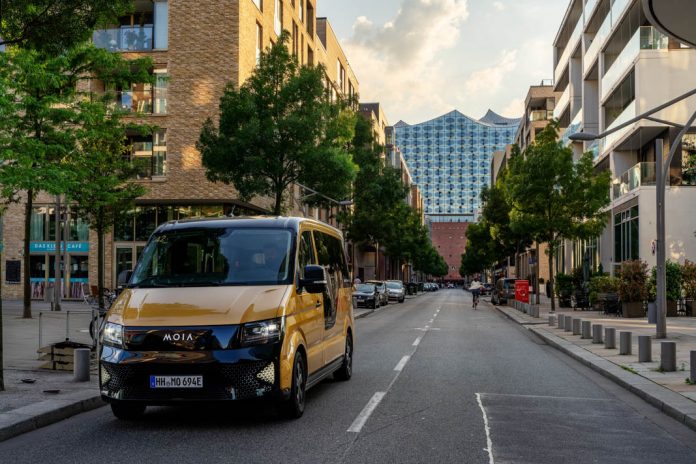Ein goldenes Moia Sammeltaxi auf einer Straße in Hamburg. Im Hintergrund sieht man die Elbphilharmononie. Foto: Moia