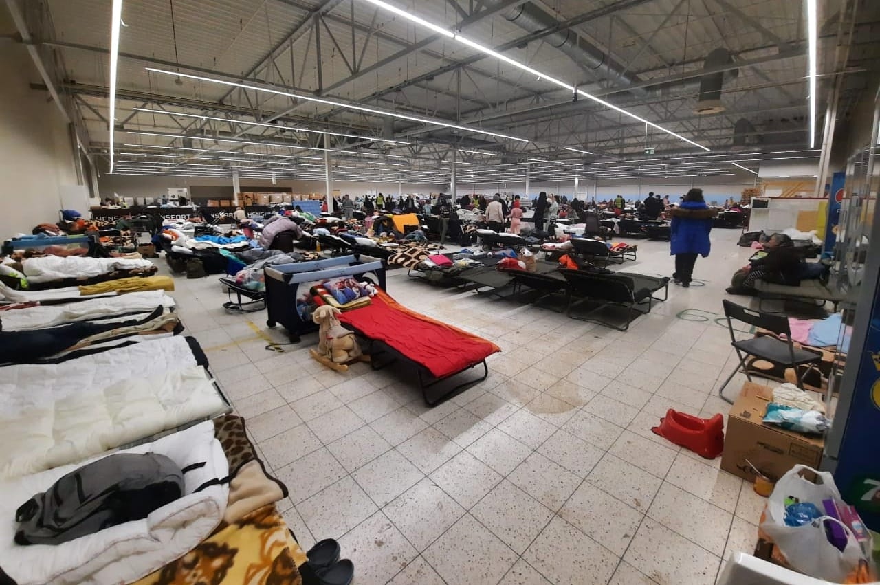 Flüchtlingslager in Przemysl von Innen. Im vorderen Teil des Bildes sind viele Betten zu sehen. Weiter hinten ist eine Menschenmenge zu sehen.