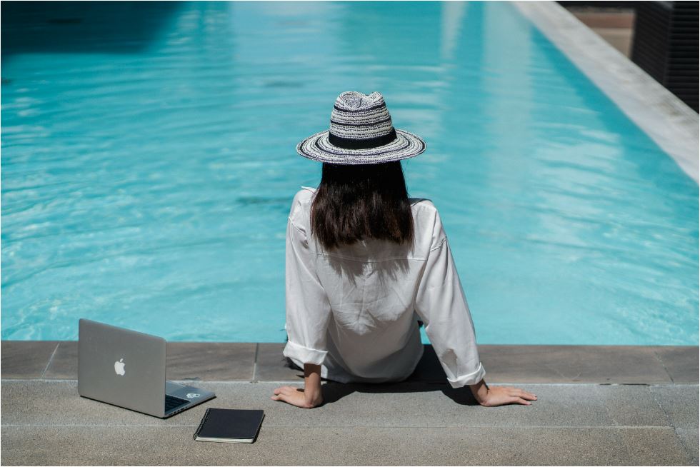 Frau sitzt neben aufgeschlagenem Laptop vor Pool in der Sonne