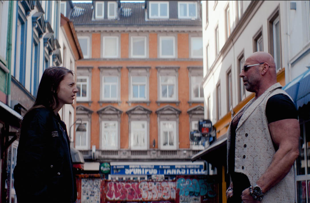 Ein Jugendlicher steht auf der linken Seite des Bildes und ein älterer Mann auf der rechten Seite. Die Straße auf der sie stehen ist auf dem Kiez in Hamburg.