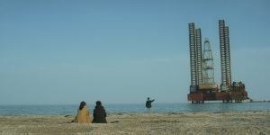 Auf dem Bild sieht man einen weitläufigen Strand. Zwei Frauen (Banu und ihre Mutter) sitzen auf dem Sand, während ein kleiner Junge (Ruslan) am Ufer näher am Wasser steht und spielt. Rechts daneben in weiterer Entfernung steht eine Ölbohrinsel.