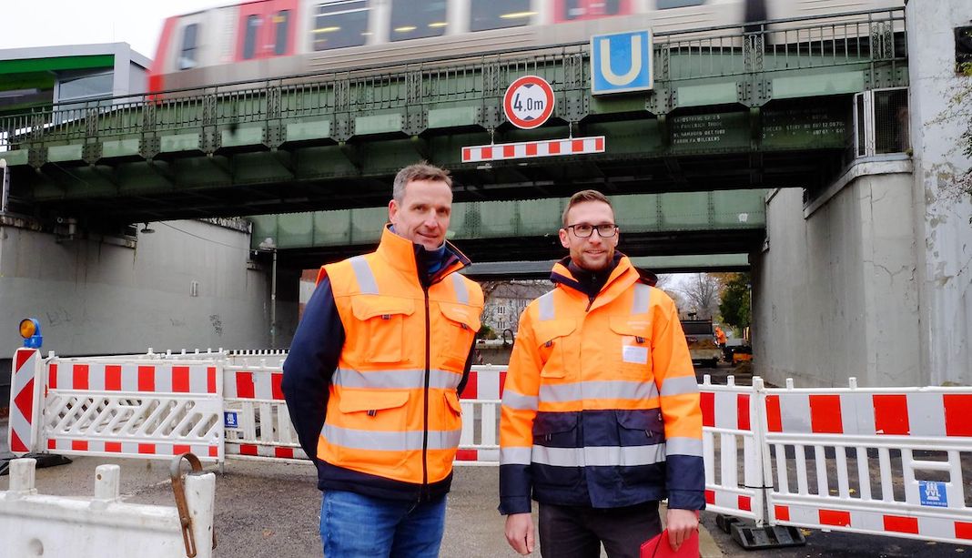 Zwei Männer mit orangenen Warnwesten vor der Hamburger U-Bahn Haltestellte Wandsbek-Gartenstadt. Dort werden vier Brücken erneuert.