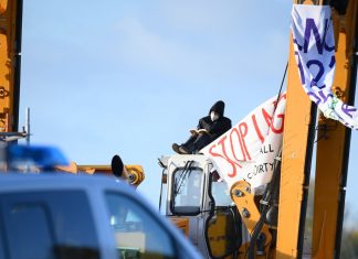 Aktivisten besetzen Baustelle für Gaspipeline Auf dem Foto sieht man ein Polizeiauto, das vor einer Baustelle parkt. Auf der Baustelle steht ein Bagger, an dem zwei große Transparente hängen. Eins davon ist lesbar: "Stop LNG" steht drauf. Auf dem Bagger sitzt ein maskierter Aktivist und liest ein Buch. Der Aktivist demonstriert gegen den Bau von Terminals für Flüssiggas.