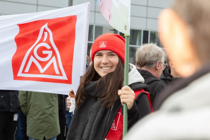 Eine Arbeitnehmerin beim Streik hält eine IG Metall-Flagge und lächelt in die Kamera. Foto: Hergen Deuter