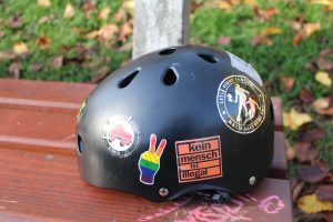 Auf dem Bild sieht man einen typischen Roller Derby Helm. Rund, schwarz und mit vielen Aufklebern. Hier zum Beispiel eine Hand, die ein Peace-Zeichen zeigt und in Regenbogenfarben ausgemalt ist. Ein anderer Aufkleber zeigt den Schriftzug "Kein Mensch ist illegal".
