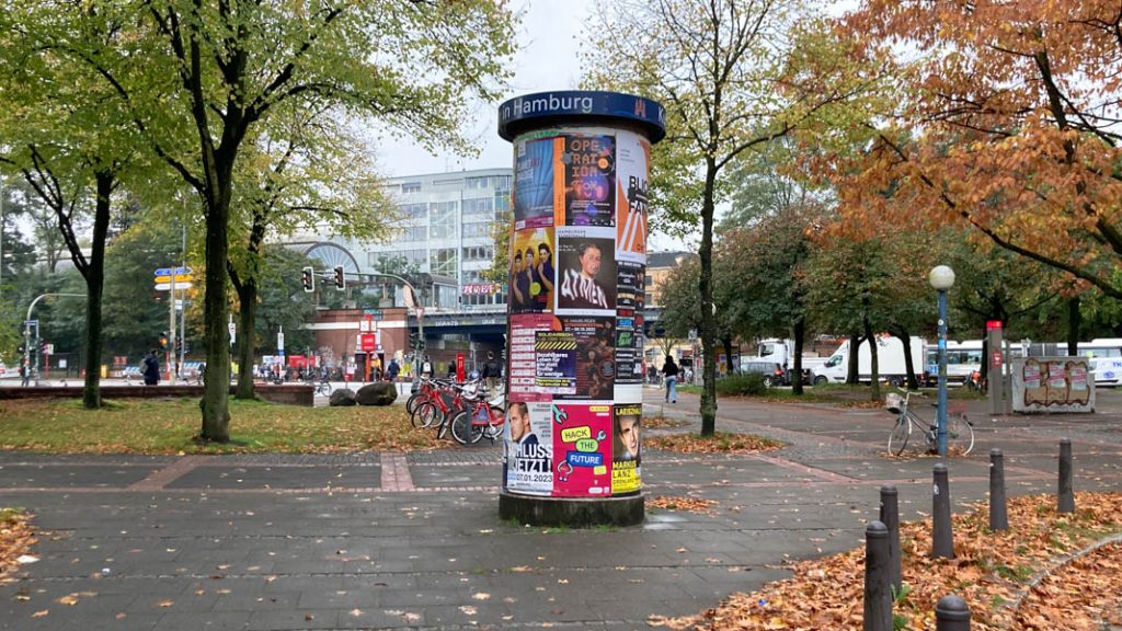 Eine analoge Litfaßsäule beklebt mit Plakaten steht zwischen Bäumen an der Holstenstraße.