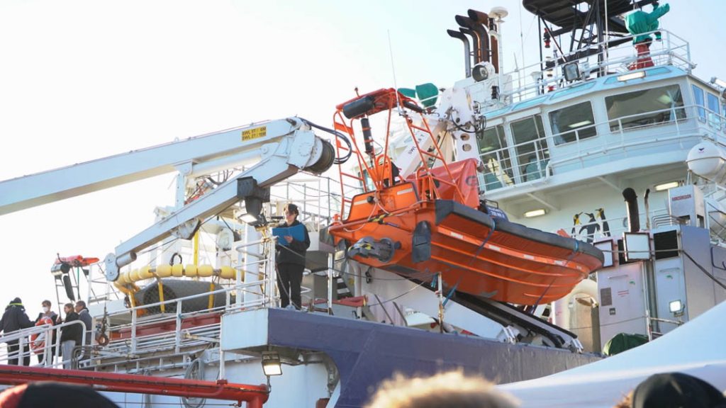 An Bord der Sea-Watch 5 steht eine Rednerin vor einem Rettungsboot