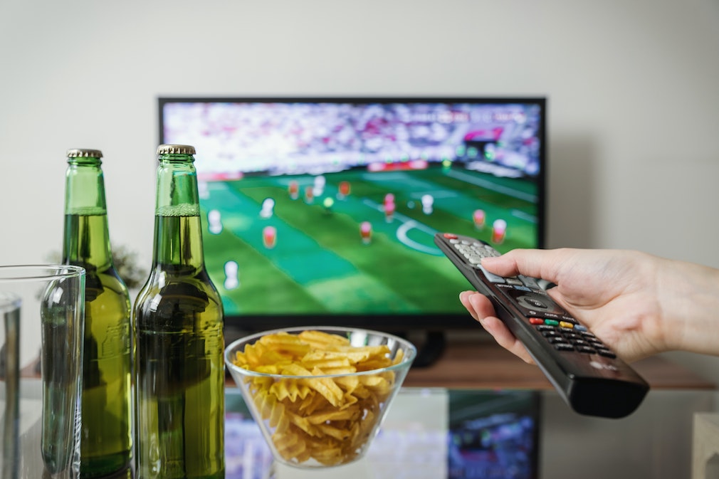 Ein Flachbildfernseher auf dem ein Fußball-Spiel läuft. Auf dem Couchtisch stehen zwei Bierflaschen und eine Schale mit Chips. ARD und ZDF zeigen Spiele der Fußball-WM in Katar.