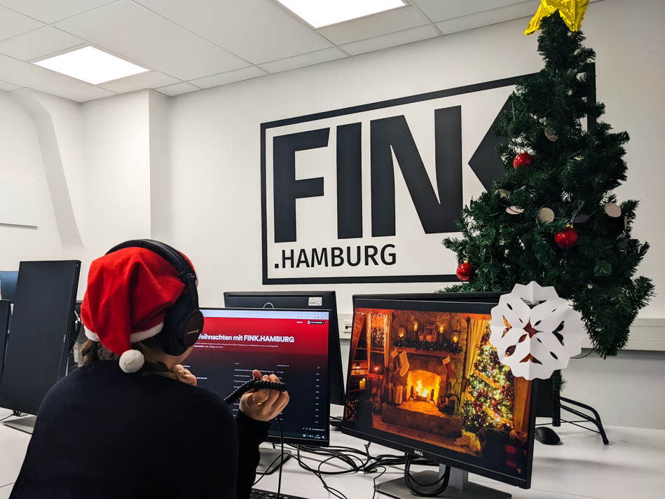 Weihnachtssongs : Lieblinge der FINK-Redaktion. Redakteurin Sarah Böse sitzt am Computer mit Kopfhörern an, im Hintergrund ein Weihnachtsbaum und das FINK.HAMBURG Logo auf einer Wand.