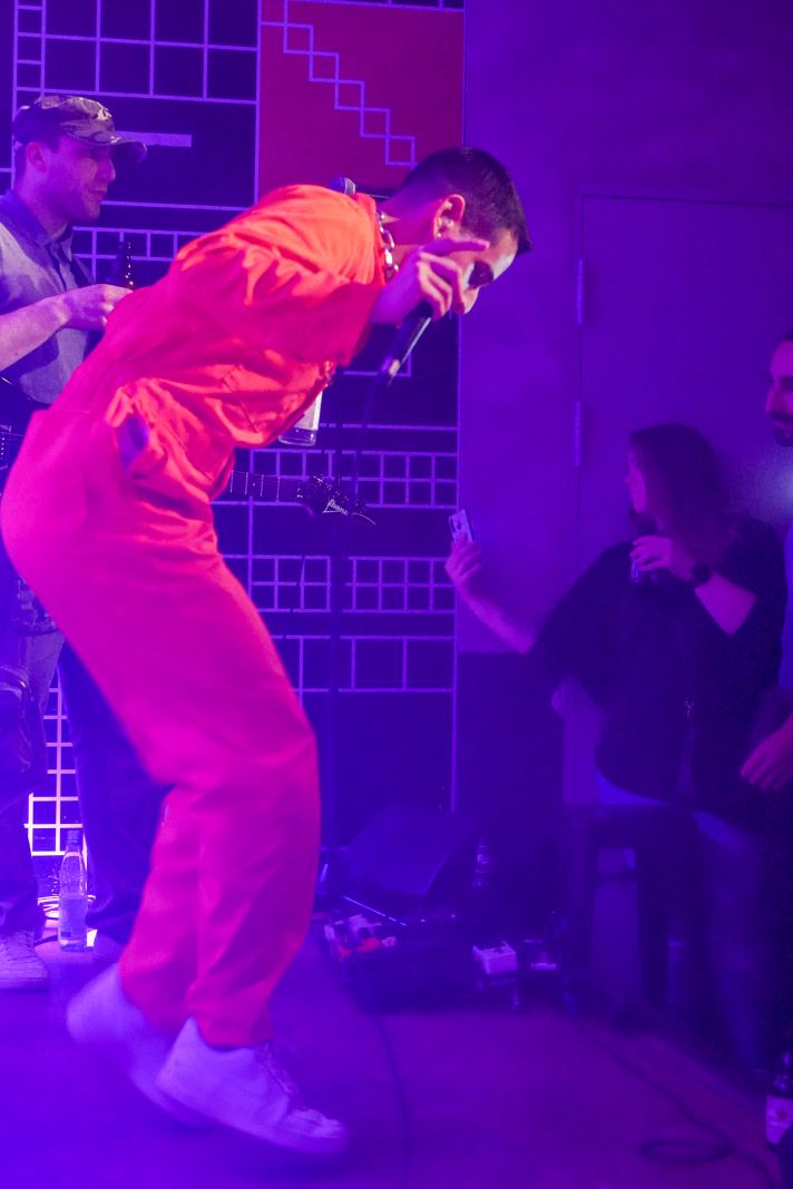 Sänger Kaso von Kaso & The A-Teem steht in einem orangefarbenen Overall auf der Bühne. Er ist in rotes Licht getaucht. Das Publikum sieht ihm aufmerksam zu.