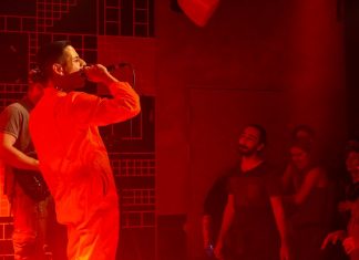 Kaso & The A-Teem: Crossover-Funk mit Flamenco Gitarre Sänger Kaso von Kaso & The A-Teem steht in einem orangefarbenen Overall auf der Bühne. Er ist in rotes Licht getaucht. Das Publikum sieht ihm aufmerksam zu.