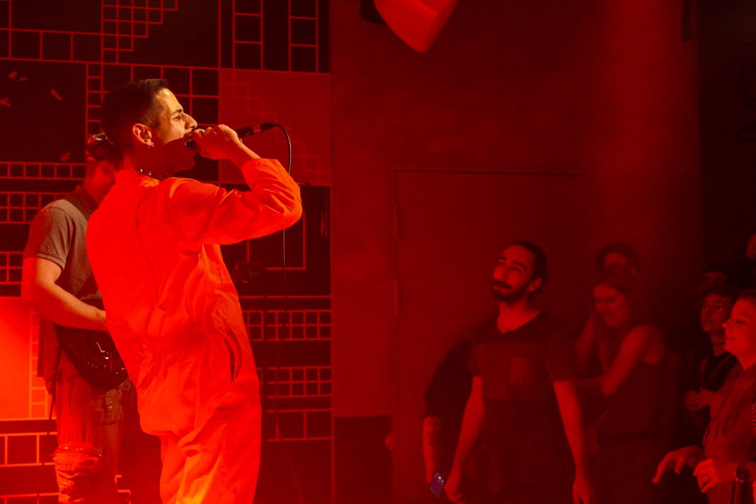 Sänger Kaso von Kaso & The A-Teem steht in einem orangefarbenen Overall auf der Bühne. Er ist in rotes Licht getaucht. Das Publikum sieht ihm aufmerksam zu.