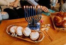 Menorah, Dreidel, Latkes: Wie feiert man Chanukkah? Auf dem Bild sieht man einen neunarmigen Kerzenleuchter, an dem alle Kerzen brennen. Daneben vier Pfannkuchen, Goldmünzen und ein Hefezopf.