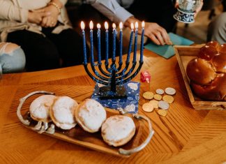 Menorah, Dreidel, Latkes: Wie feiert man Chanukkah? Auf dem Bild sieht man einen neunarmigen Kerzenleuchter, an dem alle Kerzen brennen. Daneben vier Pfannkuchen, Goldmünzen und ein Hefezopf.