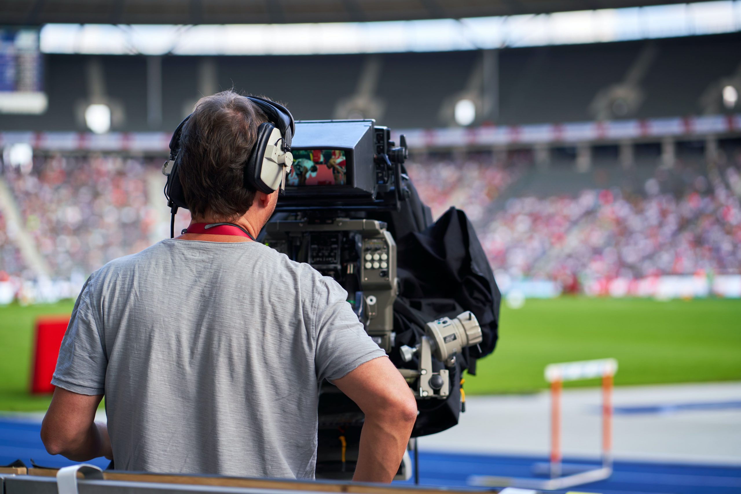 Ein Mann bedient eine TV-Kamera am Spielfeldrand eines Fußballspiels. Man sieht den Mann von hinten und guckt mit Blickrichtung der Kamera