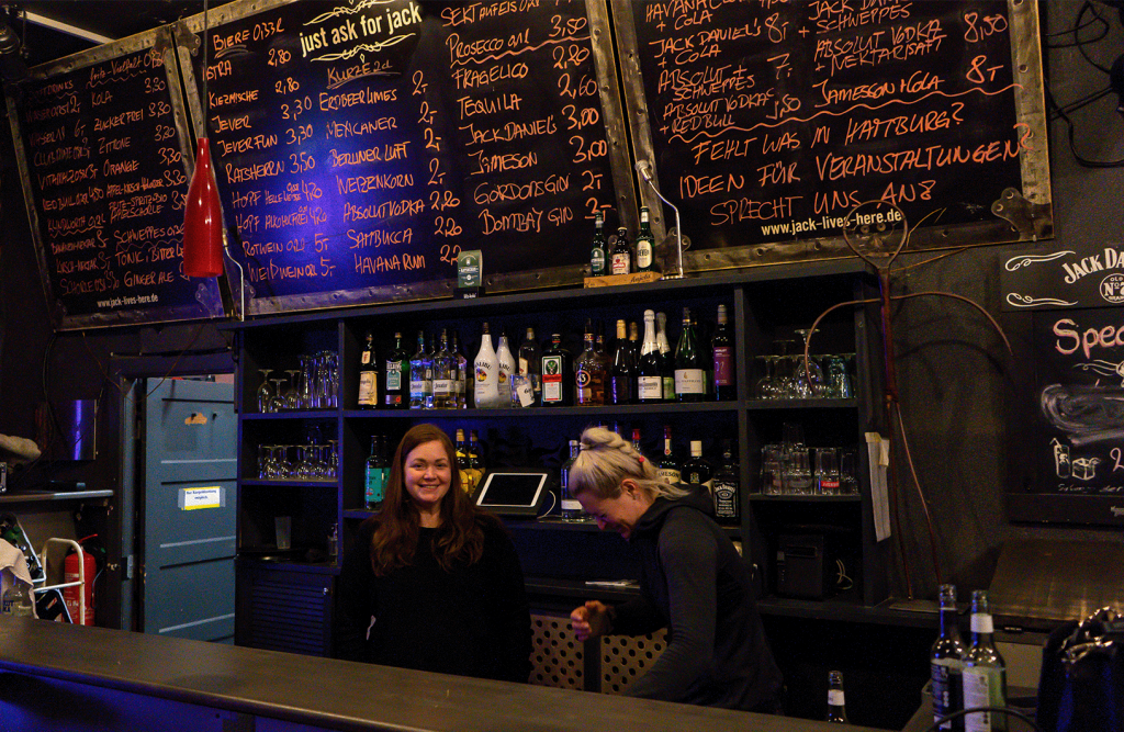 Zwei Frauen stehen an einer Bar vor Schnapsflachen, über ihnen stehen die Preise auf einer Tafel