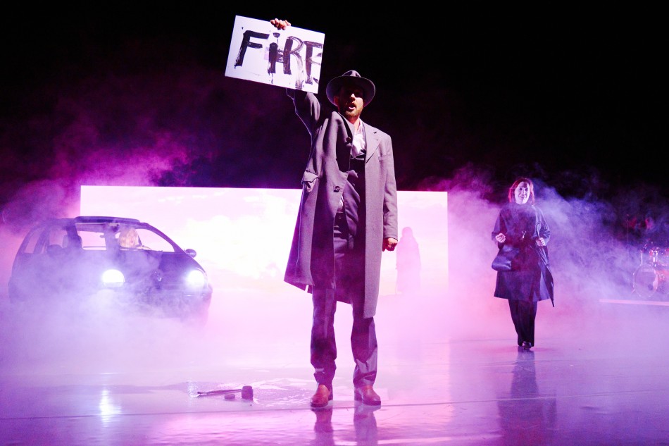 Umgeben von Nebelschwaden steht Odin Biron für die Inszenierung „Barocco” auf der Bühne. Demonstrativ hält er ein Plakat in die Luft auf dem „Fire“ steht.