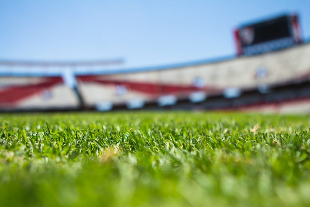 Im Vordergrund ist der Rasen eines Fußballfeldes zu sehen. Im Hintergrund ist die Tribüne eines Stadions zu erkennen.