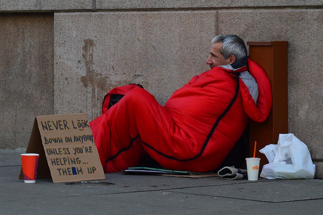 Obdachlosigkeit in Hamburg: Ein Mann liegt in einem orangenem Schlafsack auf Betonboden, neben ihm ein beschrifteter Karton und ein Getränkebecher.