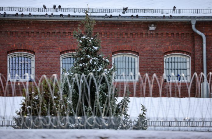 Hinter einem Stacheldrahtzaun ist ein Weihnachtsbaum zu sehen, hinter dem ein Gefängnisgebäude steht.
