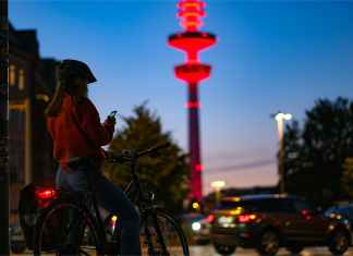 Hamburg, eine Fahrradstadt? Hamburg zur blauen Stunde, Frau mit Fahrrad im Vordergrund, dahinter der beleuchtete Fernsehturm, Straße und Bäume