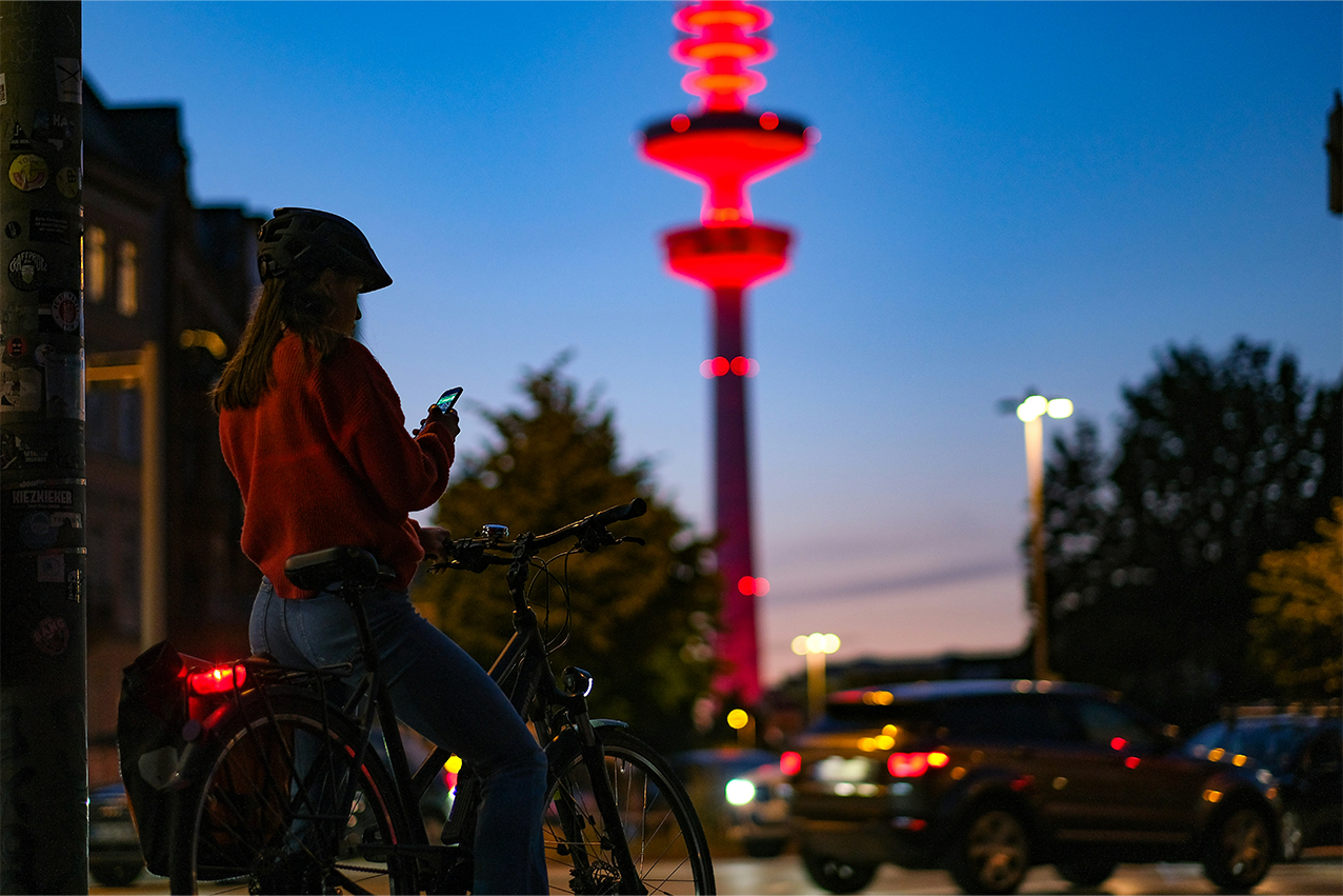 Hamburg zur blauen Stunde, Frau mit Fahrrad im Vordergrund, dahinter der beleuchtete Fernsehturm, Straße und Bäume