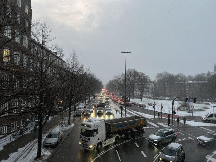 Das Bild zeigt eine Kreuzung mit vielen Autos und einem LKW. Die Bürgersteige und Grünflächen sind voller Schnee.