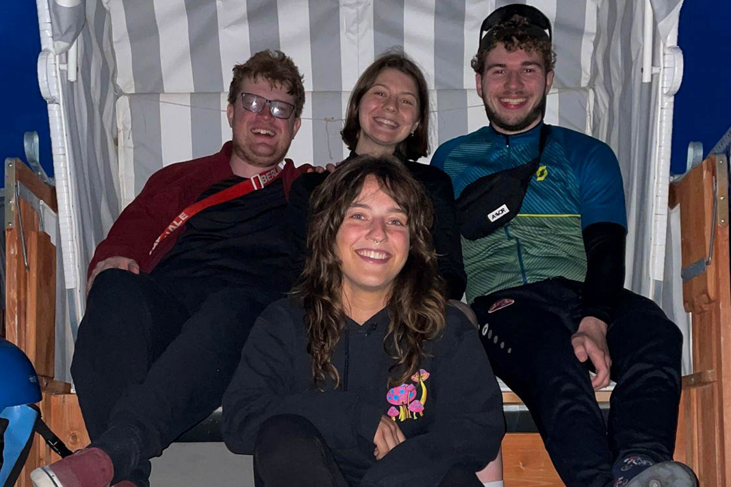 Vier junge Studierende sitzen in einem grau-weiß gestreiften Strandkorb. Sie tragen Fahrradklamotten. Im Hintergrund der dunkle Nachthimmel.