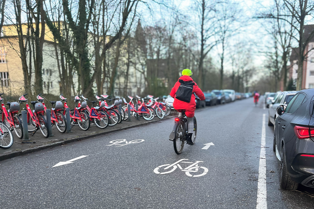 Symbolbild für Fahrradstraße in Hamburg. Eine Frau mit roter Jacke fährt auf einer Straße, auf der in beide Richtungen Piktogramme von Fahrrädern sind. Links ist eine Ausleihstation von Sharing-Fahrrädern zu sehen. Am rechten Straßenrand stehen Autos.
